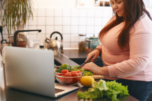 Een vrouw met een roze t-shirt staat in de keuken en snijdt groenten die op het aanrecht liggen, tegen overgewicht.