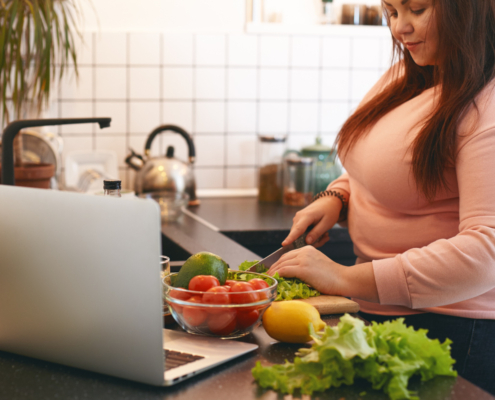 Een vrouw met een roze t-shirt staat in de keuken en snijdt groenten die op het aanrecht liggen, tegen overgewicht.