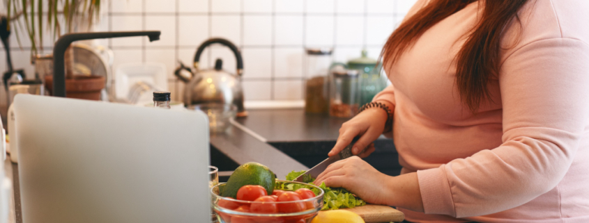 Een vrouw met een roze t-shirt staat in de keuken en snijdt groenten die op het aanrecht liggen, tegen overgewicht.
