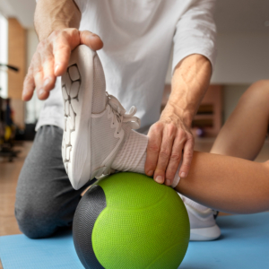 fysiotherapie voor sporters Vrouw krijgt fysiotherapie voor sporters terwijl zij last heeft van haar enkel en een oefening uitvoert op een groene bal in de oefenzaal met behulp van een therapeut in een wit shirt.